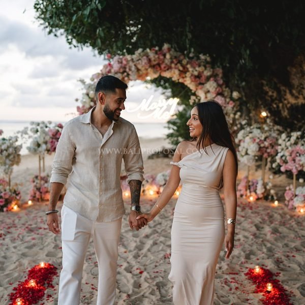 Man proposing on the beach under a bamboo triangle arch with lanterns at sunset