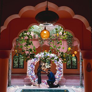 Man proposing to his partner under a flower ring in a pink arched courtyard
