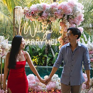 Smiling couple holding hands in front of a flower arch and a neon Will You Marry Me sign