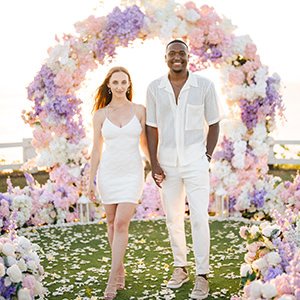 Couple walking hand in hand through a pastel flower arch at sunset